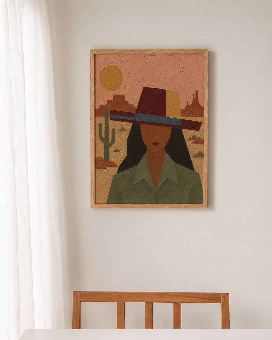 A framed art print featuring a stylized desert portrait of a woman in a wide-brimmed hat, hanging on a white wall above a wooden chair.